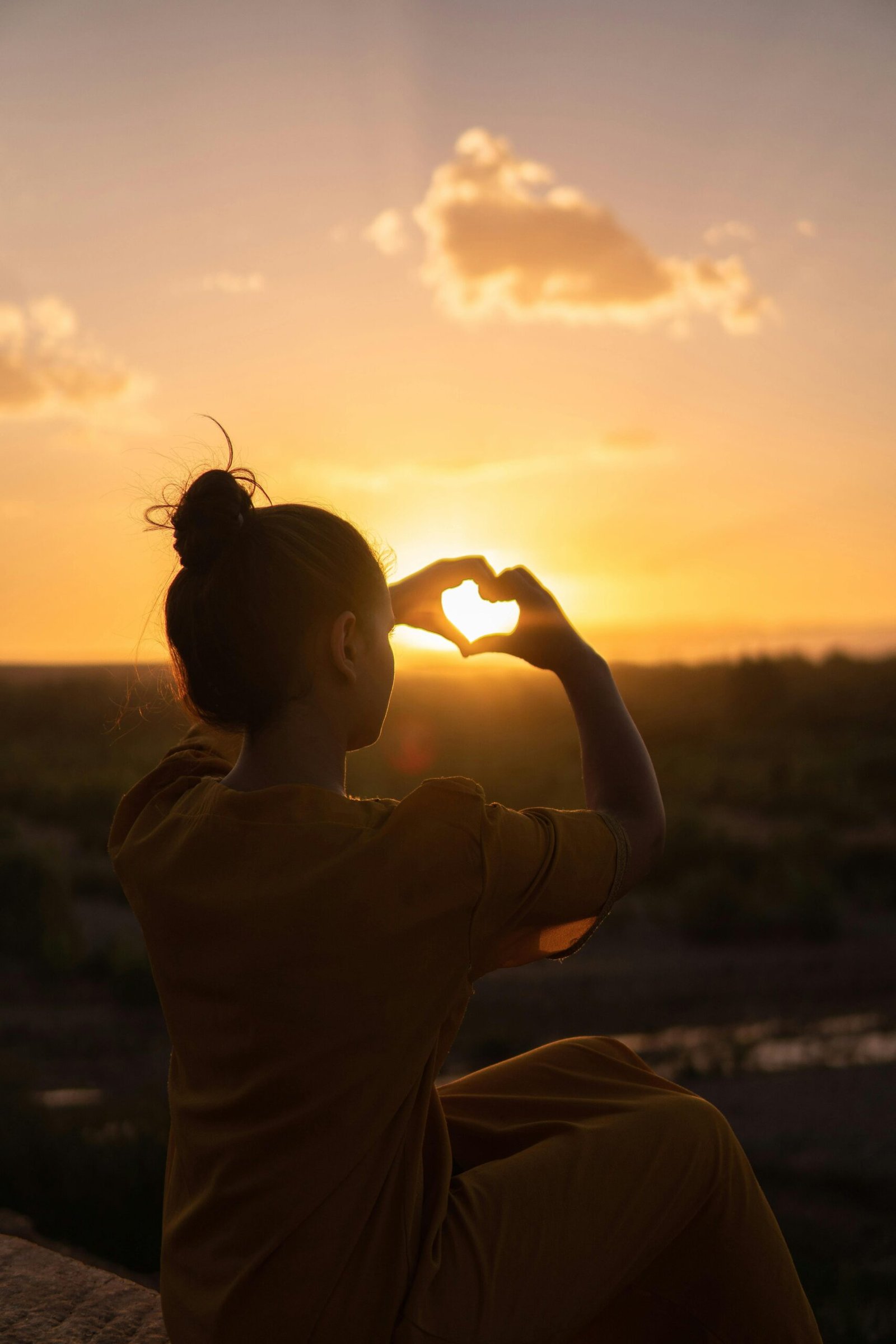 Woman standing on a cliff, with her hands in the shape of a love heart staring at a sunset